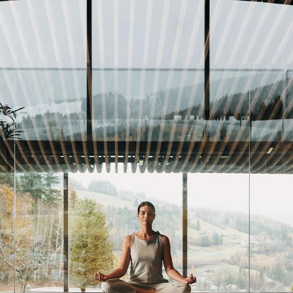 Frau meditiert im Lotussitz vor Panoramafenster mit Blick auf bewaldete Hügel