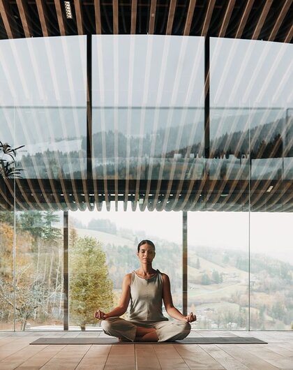 Frau meditiert im Lotussitz vor Panoramafenster mit Blick auf bewaldete Hügel