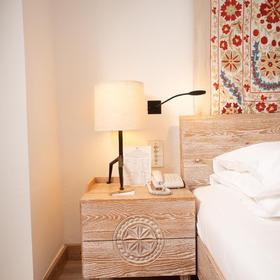 Nachttisch mit Lampe, Telefon und Dekoration neben einem Bett mit weißer Bettwäsche vor einer Wand mit Muster
