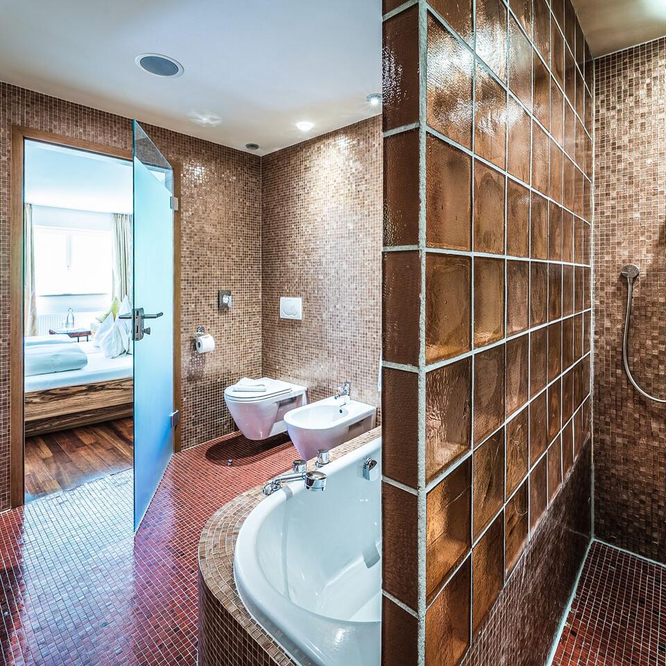 Badezimmer mit braunen Mosaikfliesen, Badewanne, Bidet, Toilette und Blick in ein Schlafzimmer