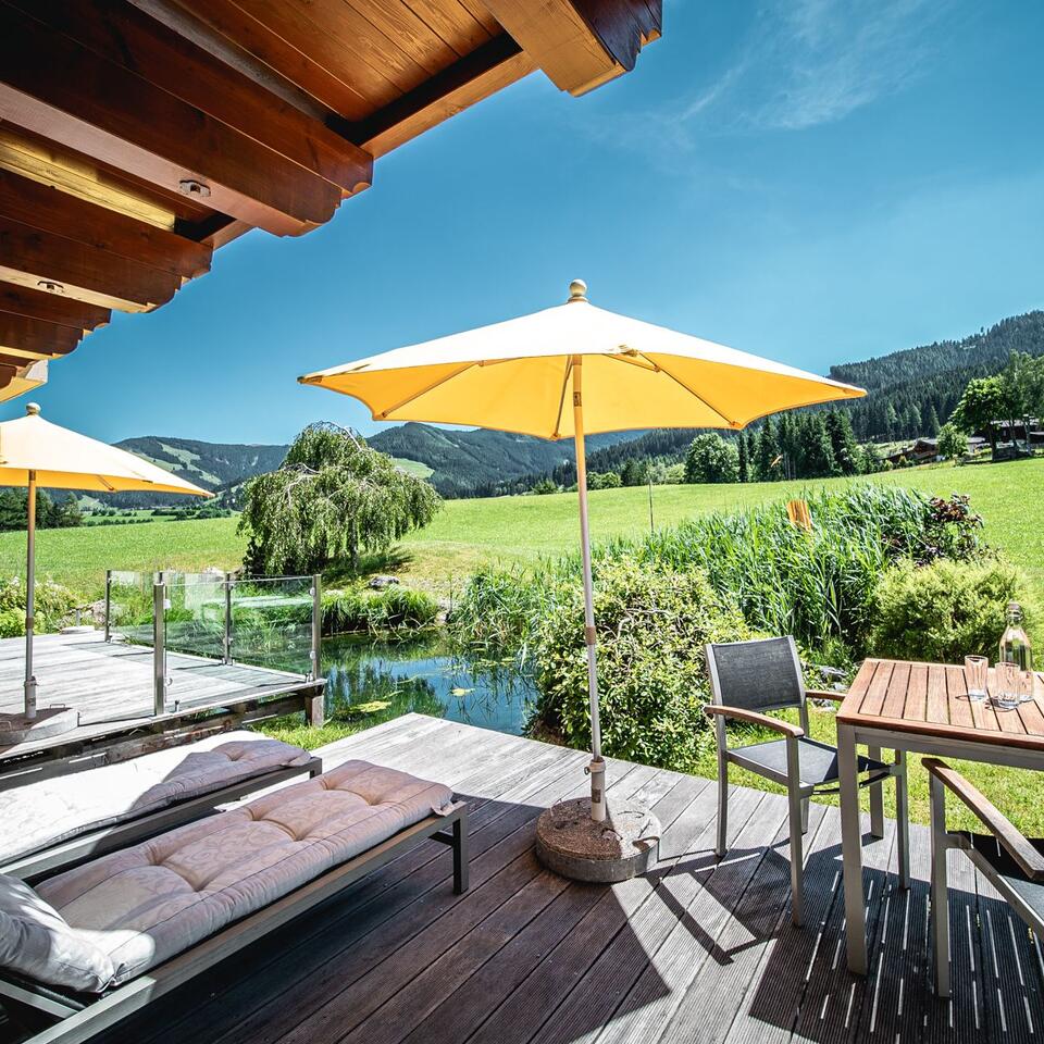 Terrasse mit Liegen, Tisch, Stühlen und Sonnenschirm vor grüner Landschaft