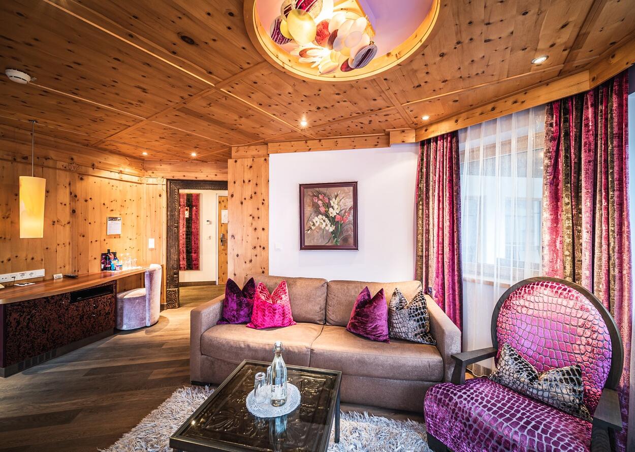 Gemütliches Hotelzimmer mit Holzwänden, Sofa, Sessel und auffälliger Deckenlampe
