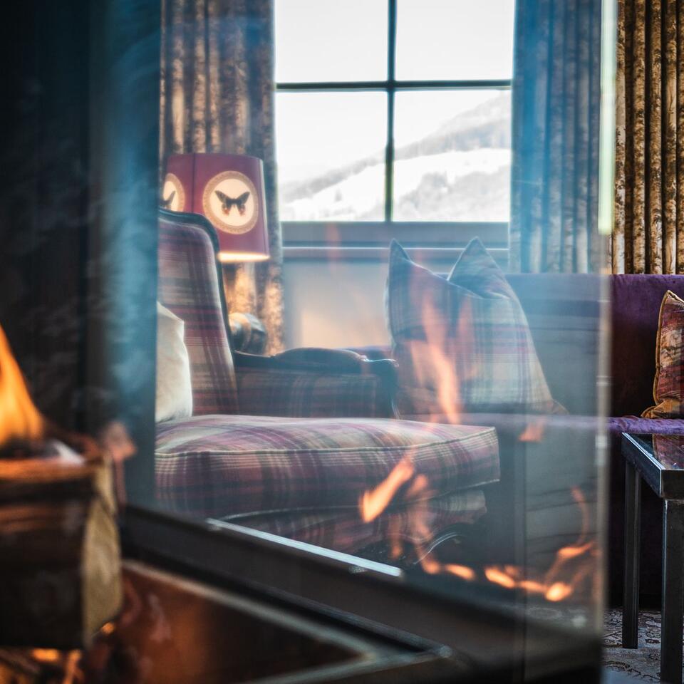 Kaminfeuer vor Wohnzimmer mit Sessel, Sofa und Fenster mit Winterlandschaft