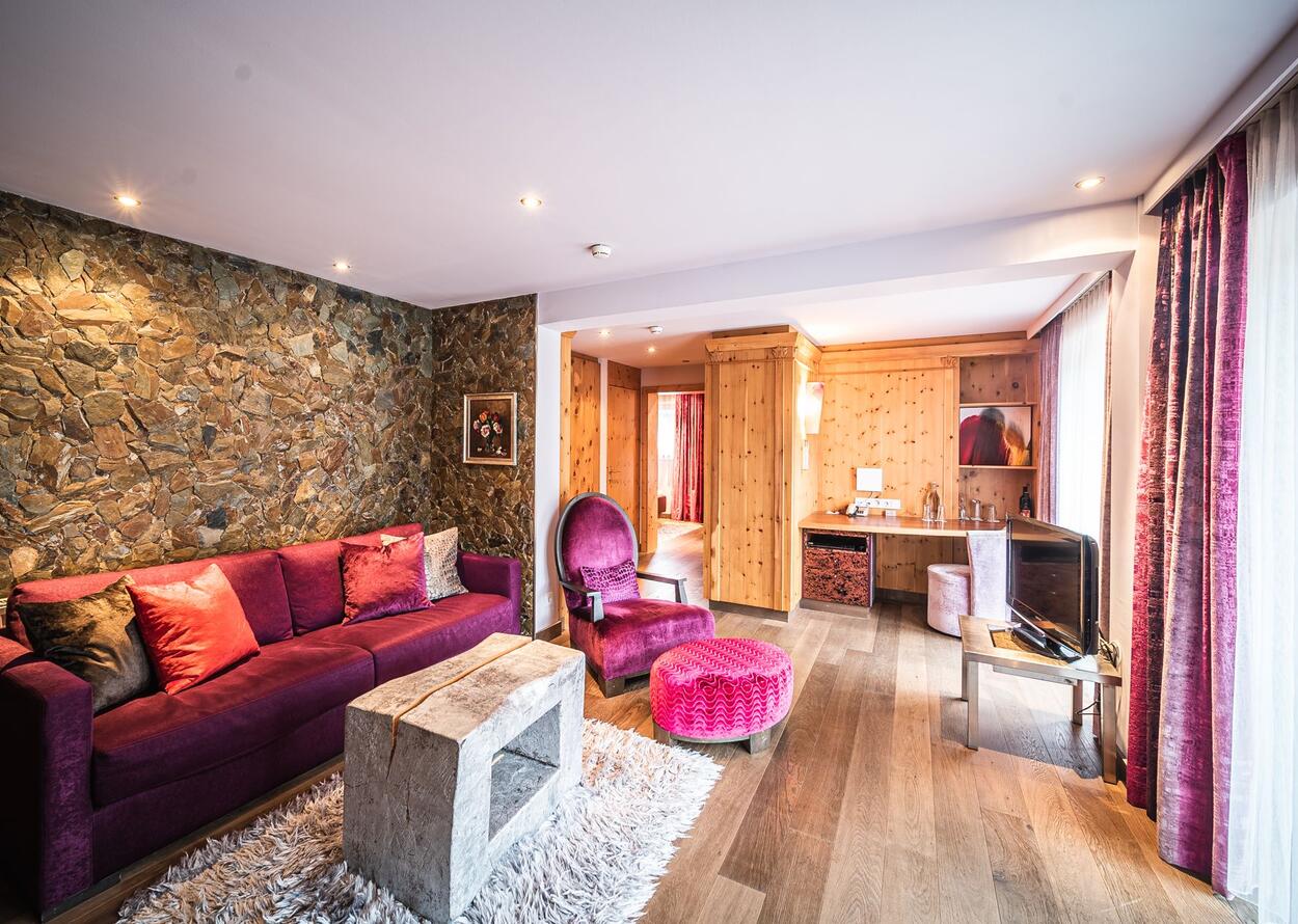 Wohnzimmer mit lila Sofa, Sessel und Hocker, Holzwand und Couchtisch aus Stein