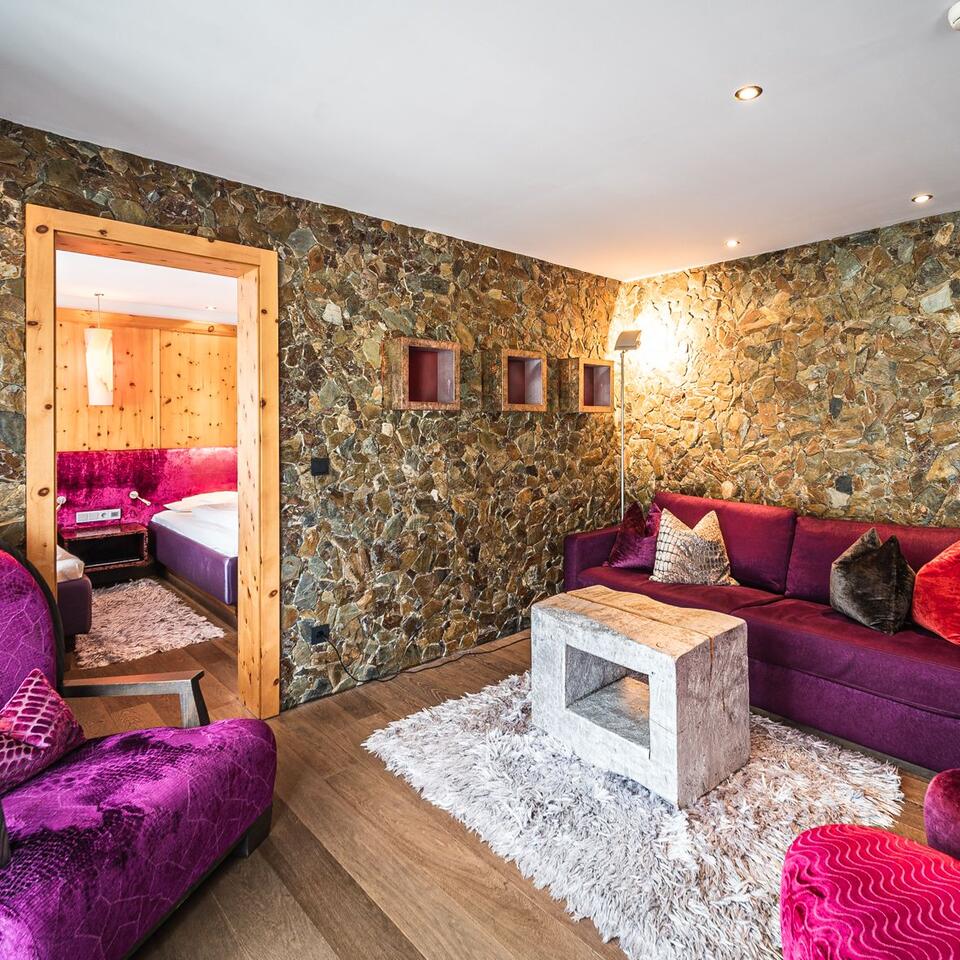 Living room with purple sofa, armchair and view into bedroom with double bed and wooden wall