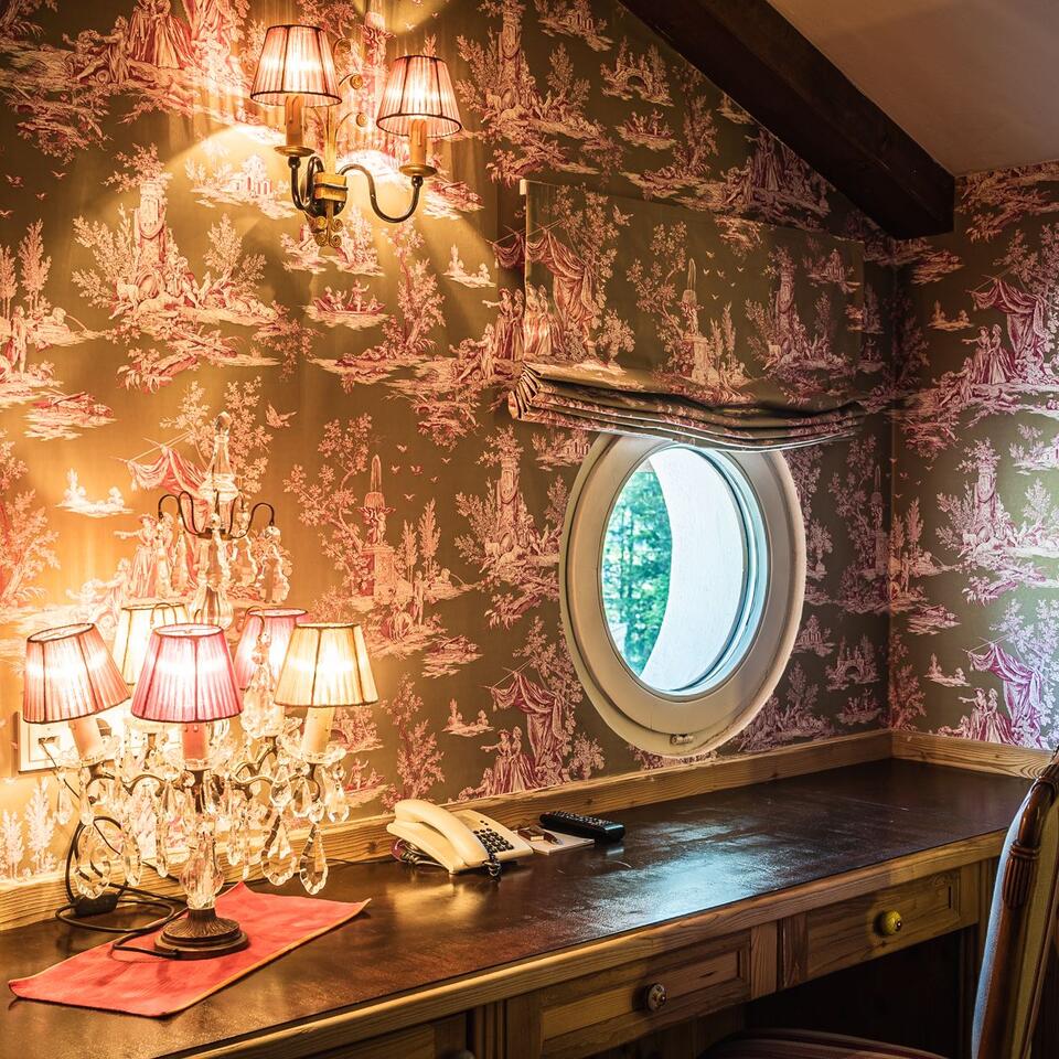 Zimmer mit Toile-de-Jouy-Tapete, rundem Fenster, Schreibtisch und Lampen