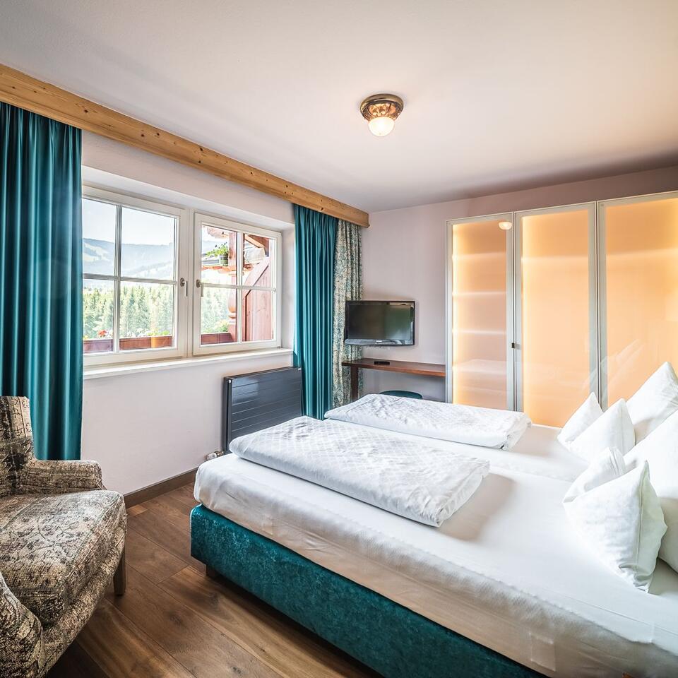 Helles Schlafzimmer mit Doppelbett, Sessel, Fenster und türkisblauen Vorhängen