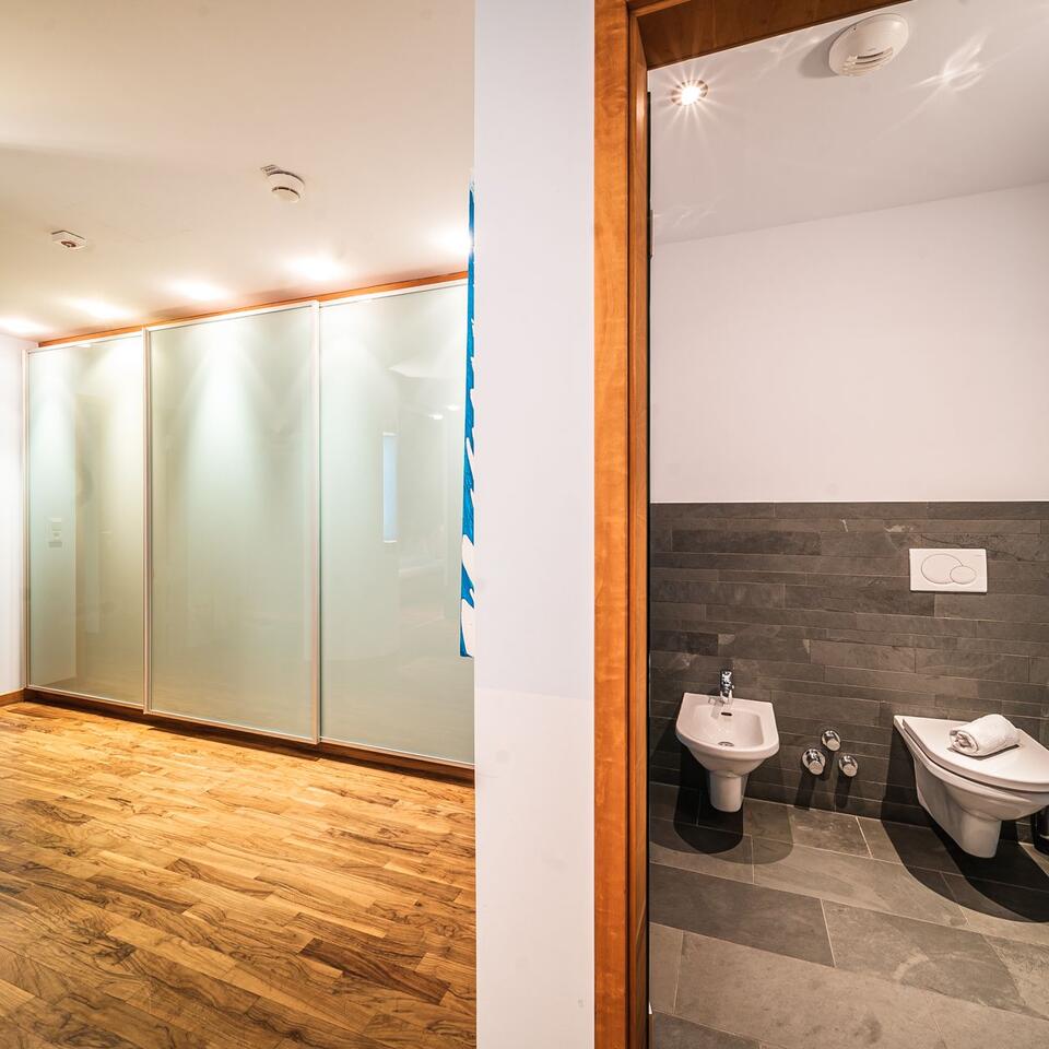 Blick in ein Badezimmer mit Toilette und Bidet neben einem Raum mit Schrankwand und Holzboden