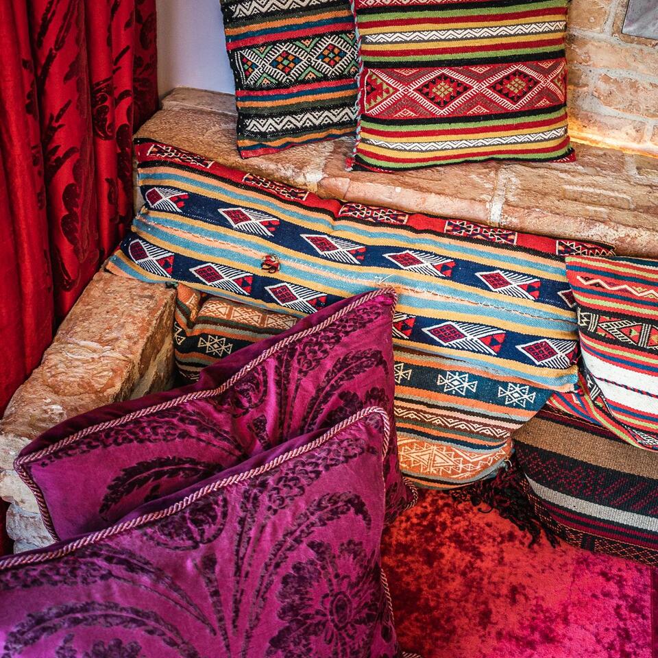 Cushions in different colors and patterns on a stone bench in front of a red curtain