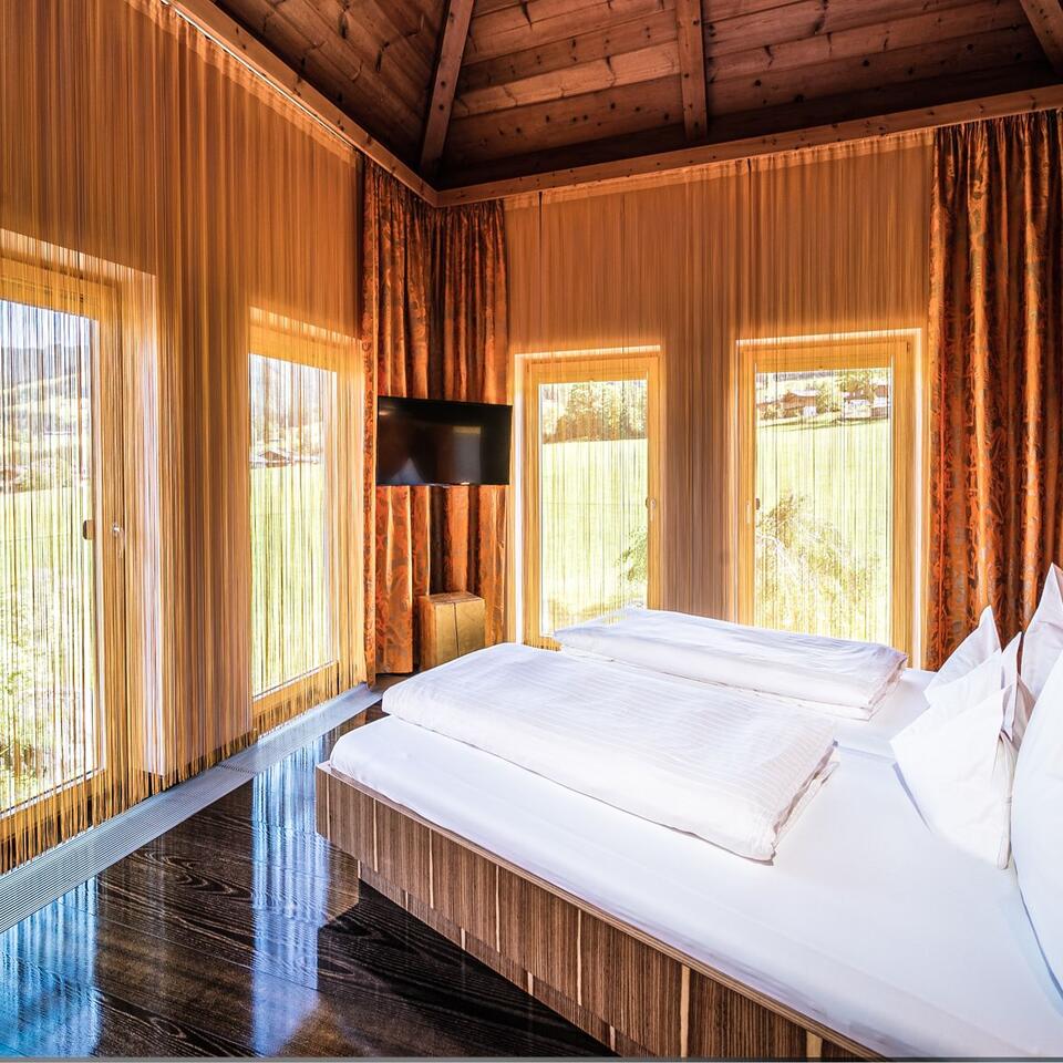 Schlafzimmer mit Holzboden, hellem Bett, Fenstern mit Vorhängen und Blick ins Grüne