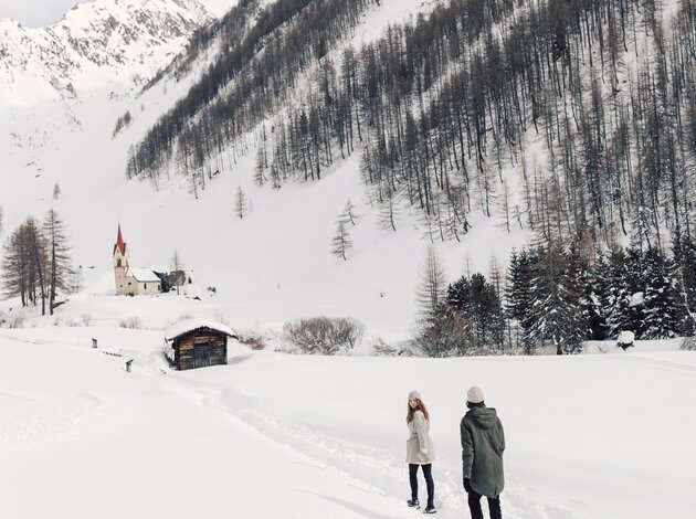 Verschneite Landschaft in Südtirol mit Kirche, Hütte und zwei Personen im Vordergrund